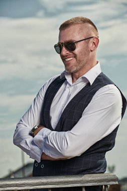 Klasik takım elbiseli ve gökyüzüne karşı modern güneş gözlüğü takmış gülümseyen olgun bir adamın portresi. Erkek tarzı. Erkekler için optik.