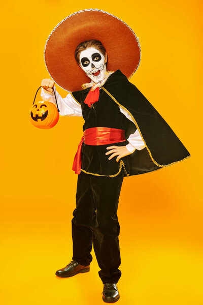 Emotional boy with sugar skull makeup trick-or-treating over bright yellow background. Halloween. Dia de los muertos. Day of the dead. Copy space.