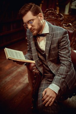 Yakışıklı beyefendi deri koltukta oturuyor ve klasik bir iç mekânda kitap okuyor. Lüks bir yaşam tarzı. Retro ve vintage tarzı. 