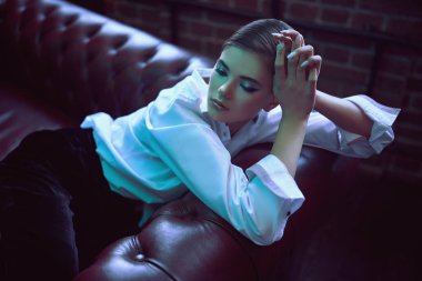 Modern genç kadın şık siyah kadife bir takım elbise ve deri bir kanepede beyaz gömlekle poz veriyor. Lüks bir yaşam tarzı. Moda, makyaj ve güzellik trendleri.