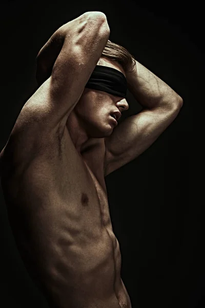 Art portrait of a handsome blindfold young man with perfect muscular body looking up at the light. Black background, darkness.