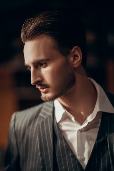 Portrait of a handsome brunet man in an elegant suit standing in a dark room. Male beauty, fashion.