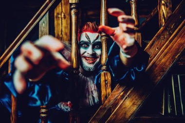 Merdiven parmaklıklarından elini çeken gülümseyen bir palyaço portresi. Cadılar Bayramı, karnaval.