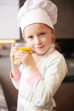 Önlük giymiş küçük tatlı bir kız portresi ve bir bardak portakal suyuyla şef şapkası. Taze meyve suyu ve sağlıklı yiyecekler..