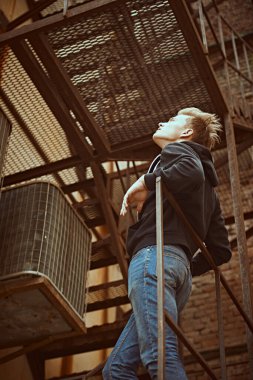 Günlük giysiler içindeki yakışıklı genç adam eski demir bir merdivende duruyor. Şehirli grunge stili. Erkek modası.