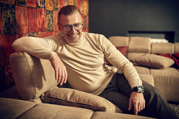 Handsome middle-aged man in elegant casual clothes and glasses having a rest and reading magazine  at home. 