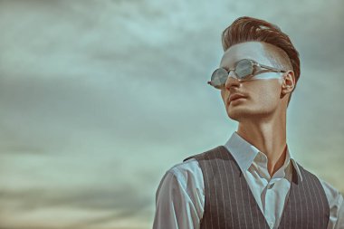 Erkek moda konsepti. Yüzünde beyaz boya olan yakışıklı bir adamın sanat portresi zarif bir takım elbise ve bulutlu gökyüzüne karşı güneş gözlüğü takıyor. Vücut boyama. Makyaj ve kozmetik.