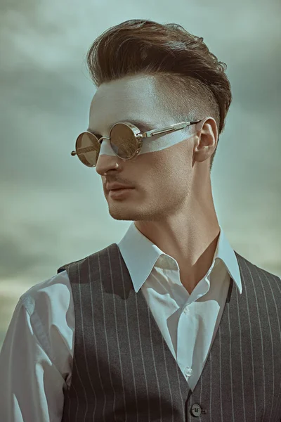 Men's fashion concept. Art portrait of a handsome man with white paint on his face posing in elegant suit and sunglasses against the cloudy sky. Body painting. Makeup and cosmetics.