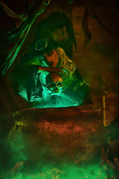 Horrible old witch holding the skull above the boiling potion in a cauldron surrounded by a magical green light. Halloween. Witchcraft.