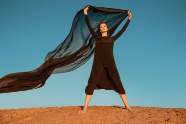 Moda fotoğrafı. Çölde poz veren uzun siyah elbiseli güzel esmer kadın rüzgarda çırpınan bir eşarpla. Gökyüzü arkaplanı.
