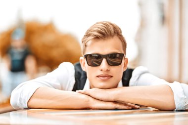 Kafenin verandasında poz veren yakışıklı bir genç adamın portresi. Sonbahar modası. Erkekler için optik.