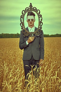 Erkek moda konsepti. Zarif takım elbiseli yakışıklı bir adamın portresi ve modaya uygun saç stili buğday tarlasında klasik resim çerçevesiyle poz veriyor. Vücut boyası. 