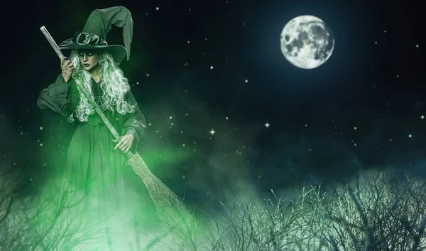 A scary ugly witch standswith a broom in the forest against the background of the starry sky and the moon. Halloween. Scary tales.