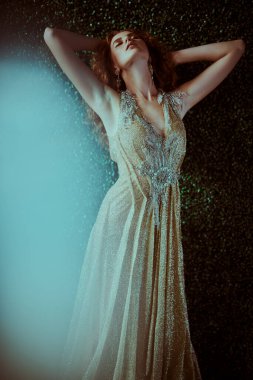 Koyu yeşil bitki arka planında lüks bir altın elbiseyle poz veren güzel kızıl saçlı bir kadının portresi. Güzellik, akşam modası.. 