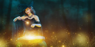 Cadılar Bayramı. Şapkalı genç ve güzel bir cadı ve sihirli bir değnekle karanlık bir ormanda sihirli ışıklarla çevrili bir büyü yapıyor. Boşluğu kopyala. 