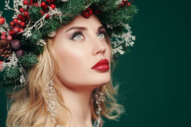 Kafasında Noel çelengi olan, Noel topları, böğürtlenler ve kar taneleriyle süslenmiş güzel, mutlu genç bir kadın. Yeşil arka plan. Boşluğu kopyala.