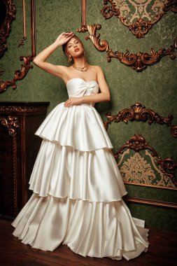 Lüks bir dairede gösterişli beyaz elbiseli ve değerli mücevherlerle poz veren muhteşem bir genç kadın. Klasik klasik iç mimari. Düğün modası.