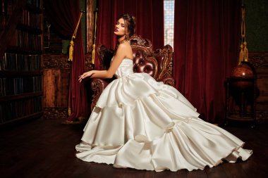 Lüks beyaz elbiseli ve değerli mücevherleri olan muhteşem genç bir kadın lüks bir dairede bir sandalyede oturuyor. Klasik klasik iç mimari. Düğün modası.