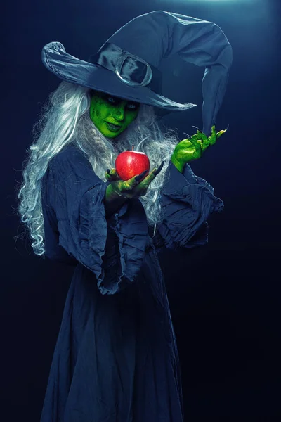 Halloween. Scary green old witch holds out a red apple. Woodland background, evil black magic.