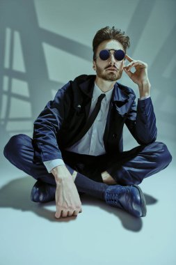 Zarif siyah takım elbiseli, beyaz gömlekli ve güneş gözlüklü, gri arka planda, pencere gölgeli modern tarz bir adam. Erkek modası.