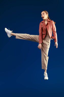 Heyecanlı, pantolonlu, deri ceketli, geniş bir adım atıp koyu mavi arka planda ağzını açan modern bir kız modelin portresi. Güzellik, moda. Boşluğu kopyala. 