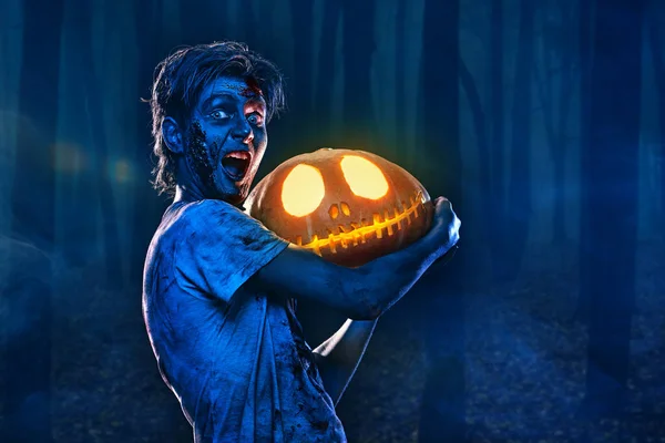 Halloween concept. Portrait of a scary zombie boy teenager holding a pumpkin lantern in forest. Copy space.