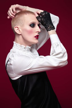 Kadın makyajlı ve kadın kıyafetleriyle stüdyoda ifade ile poz veren genç bir adamın tam boy portresi..