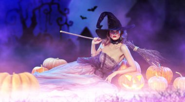 Büyüleyici güzel cadı, elinde balkabağı feneri ve lüks bir gece elbisesi ve şapkası olan bir cadı süpürgesi ile yatıyor. Cadılar Bayramı kutlaması, parti. Uzayı kopyala Cadılar Bayramı modası.