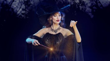 Büyüleyici, şapkalı genç bir cadının portresi ve sihirli bir değnekle sihirli ışıklarla çevrili gece ormanının arka planında. Cadılar Bayramı kutlaması, parti. 