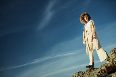 Kıvırcık saçlı, gökyüzü arka planında şık kıyafetler giyen çekici bir genç kız. Moda, güzellik.