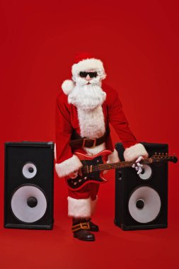 Noel Baba rock müzisyeni büyük hoparlörlerin arka planında elektro gitar çalar. Kırmızı şenlik arka planı. Noel ve Yeni Yıl Partisi. 