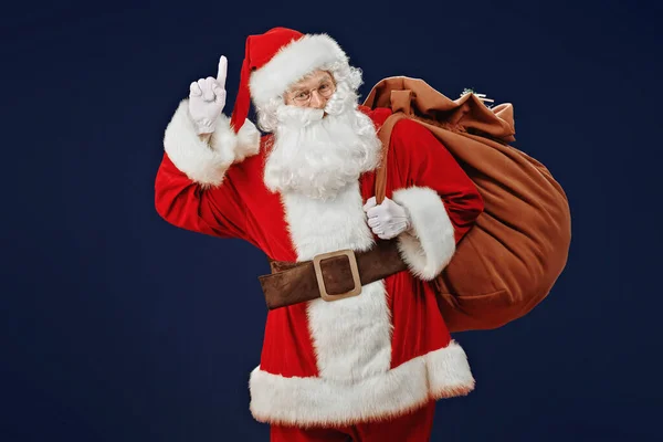 Attention, Christmas! Santa Claus raises his index finger to attract attention. Studio portrait over dark blue background.