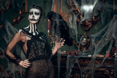 Kafatası kabağı makyajlı zarif bayan eski kale dekorasyonunda siyah kuzgun duruyor. Eski tarz. Cadılar Bayramı. Ölülerin Günü. 