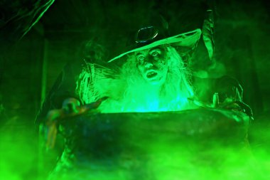 Korkunç yaşlı cadı, sihirli yeşil ışıkla çevrili kazanında iksir yapıyor. Cadılar Bayramı. Korkunç hikayeler..