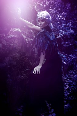 Güzel gri saçlı ve zengin siyah elbiseli bir cadı sihirli ışıkla ormanda poz veriyor. Gizemciliğin atmosferi. Cadılar Bayramı.