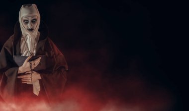 Elinde İncil ve elinde haç olan lanetli bir rahibe, kan kırmızısı bir sis ile siyah bir arkaplanda kameraya bakıyor. Cadılar Bayramı. Dehşet. Boşluğu kopyala.