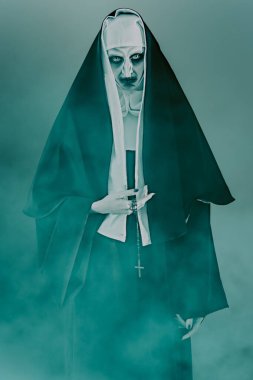 Korkunç şeytani ele geçirilmiş rahibe elinde haçla karanlık bir odada duman içinde duruyor. Korku ve Cadılar Bayramı.