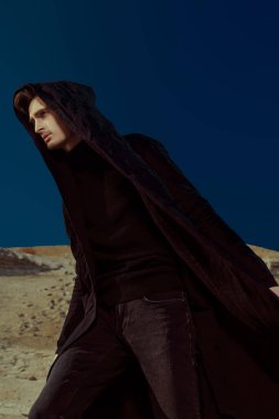 Siyah giysiler içinde çölde yürüyen yakışıklı bir esmer adamın portresi. Kumlar ve açık mavi gökyüzü. Erkek modası.
