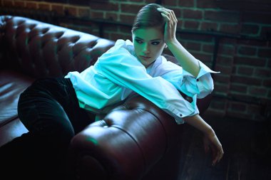 Modern genç kadın şık siyah kadife bir takım elbise ve deri bir kanepede beyaz gömlekle poz veriyor. Lüks bir yaşam tarzı. Moda, makyaj ve güzellik trendleri.