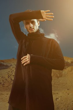Çöl. Siyah elbiseli yakışıklı bir esmer adam portresi güneşin yakıcı ışınlarının altında çölde duruyor. Erkek güzelliği, moda.