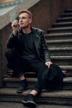 Modern genç hippi adam merdivenlerde oturuyor ve sokakta sigara içiyor. Tam boy portre. Sokak tarzı.
