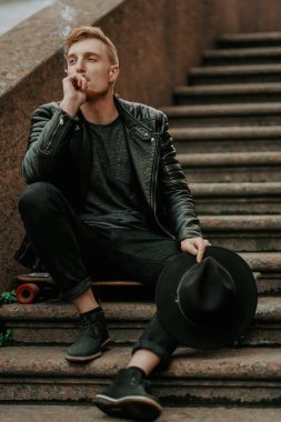 Modern genç hippi adam merdivenlerde oturuyor ve sokakta sigara içiyor. Tam boy portre. Sokak tarzı.