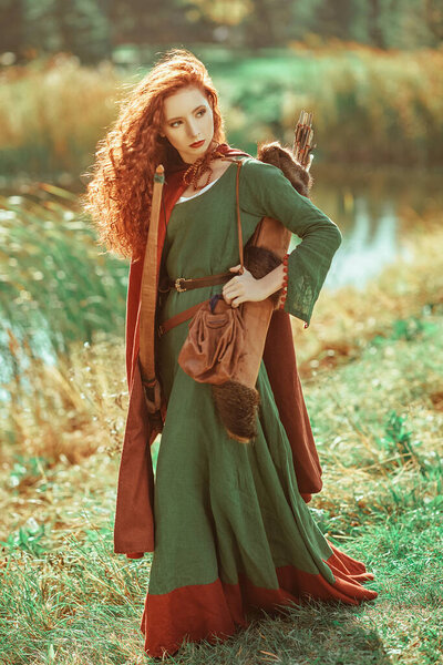 A beautiful red-haired girl archer of the Middle Ages stands next to the lake. Celtic culture. Fantasy world.