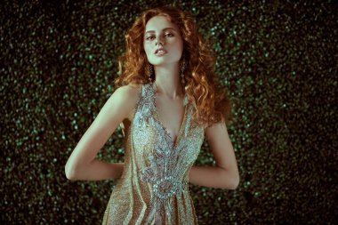 Koyu yeşil bitki arka planında lüks bir altın elbiseyle poz veren güzel kızıl saçlı bir kadının portresi. Güzellik, akşam modası.. 