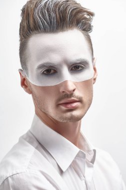 Yüzünde beyaz boya ve beyaz gömlekle beyaz duvarda poz veren yakışıklı bir adamın moda portresi. Erkek güzeli, moda. Saç stili ve kozmetik. 