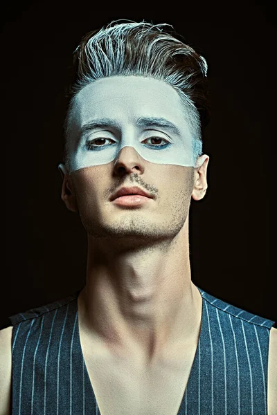 Art portrait of a handsome young man with white paint on his face on a black background with light. Male beauty, fashion. Hairstyle and cosmetics.