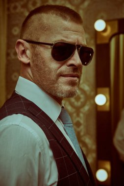 Lüks bir dairede duran şık takım elbiseli, olgun, yakışıklı bir adamın portresi. Erkek güzeli, moda. Optik, erkekler için güneş gözlüğü..