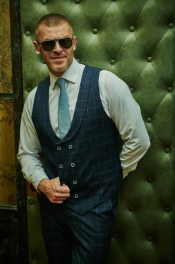 Lüks bir dairede duran şık takım elbiseli, olgun, yakışıklı bir adamın portresi. Erkek güzeli, moda. Optik, erkekler için güneş gözlüğü..