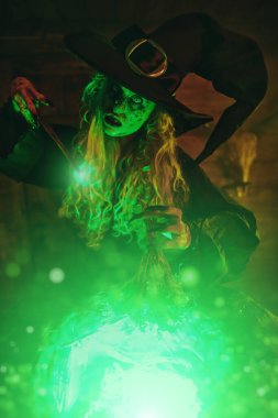 Korkunç yaşlı cadı sihirli yeşil ışıkla çevrili sihirli bir değnekle kazanda iksir yapıyor. Cadılar Bayramı. Korkunç hikayeler..