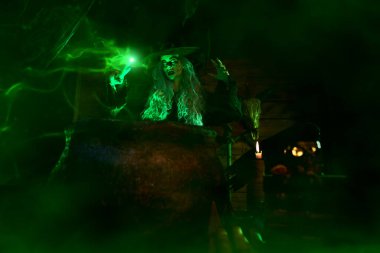 Korkunç yaşlı cadı, sihirli yeşil ışıkla çevrili kazanın yanında bir büyü yapıyor. Cadılar Bayramı. Korkunç hikayeler..